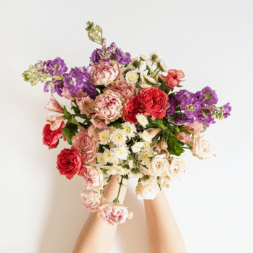 Zwei Hände halten einen bunten Blumenstrauß mit roten, rosa, weißen und violetten Blüten vor einem hellen, neutralen Hintergrund.